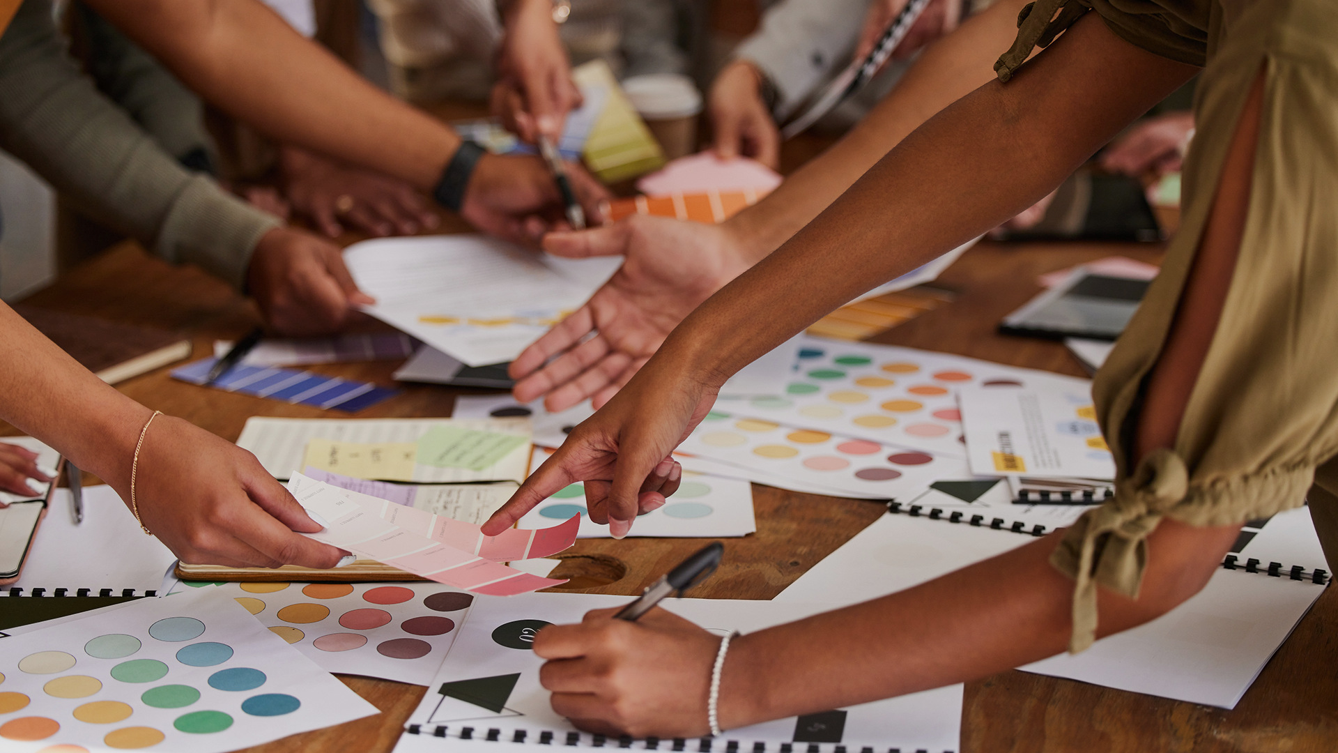 Group of team members reaching over a table filled with colour swatches, notebooks, and printed materials during a collaborative design session.